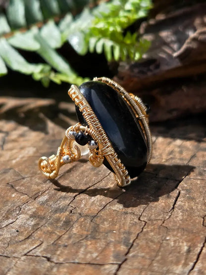 A gold copper wire-wrapped ring featuring a large, polished black oval stone, displayed on a textured wooden surface with green fern leaves in the background.