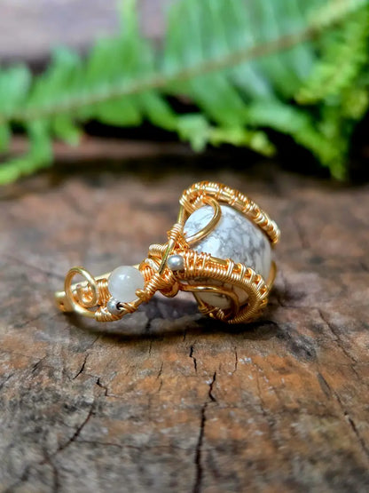 A handmade copper wire-wrapped gemstone pendant featuring a white howlite stone, resting on a textured wooden surface with a blurred green fern in the background.