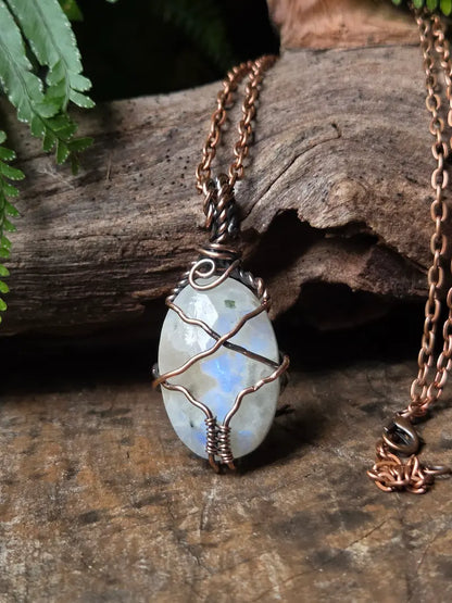 A handmade oval pendant necklace featuring a light blue-gray iridescent moonstone wrapped in copper wire, hanging from a copper chain on a rustic wood background.