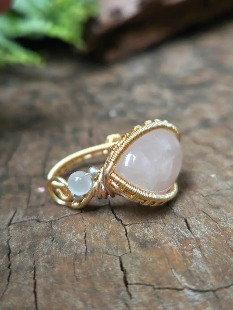 A handmade gold copper wire-wrapped ring featuring a rose quartz pink crystal and a small round white accent bead, resting on a natural wooden surface.