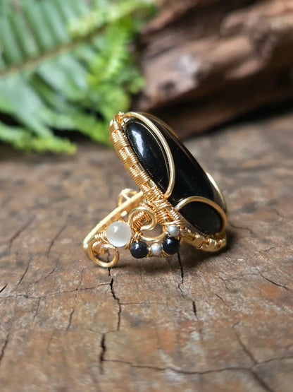 A cooper wire-wrapped ring featuring a large black stone and small white and black beads, displayed on a textured wooden surface with a blurred fern in the background.