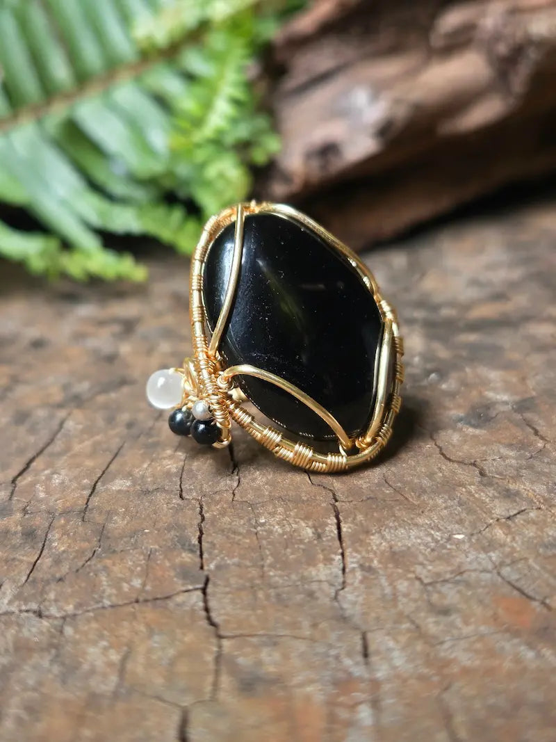 A handmade wire-wrapped ring featuring a polished black stone, set against a rustic wood background with a blurred green fern leaf.