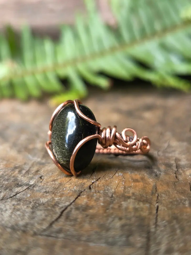 A handmade copper wire ring featuring a black Obsidian stone, resting on a textured wooden surface with a blurred green fern in the background.