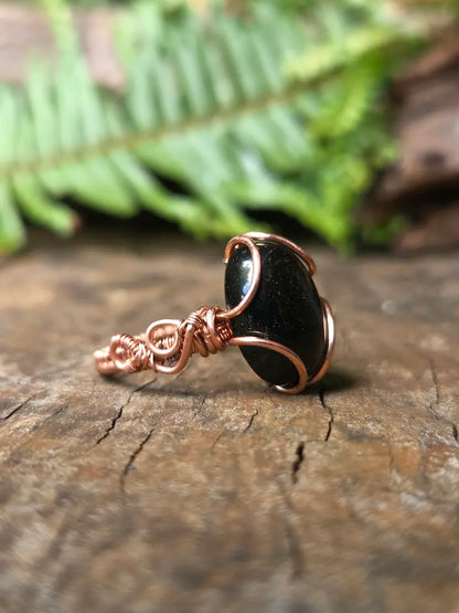 A handmade copper wire ring featuring a black Obsidian stone, resting on a textured wooden surface with a blurred green fern in the background.