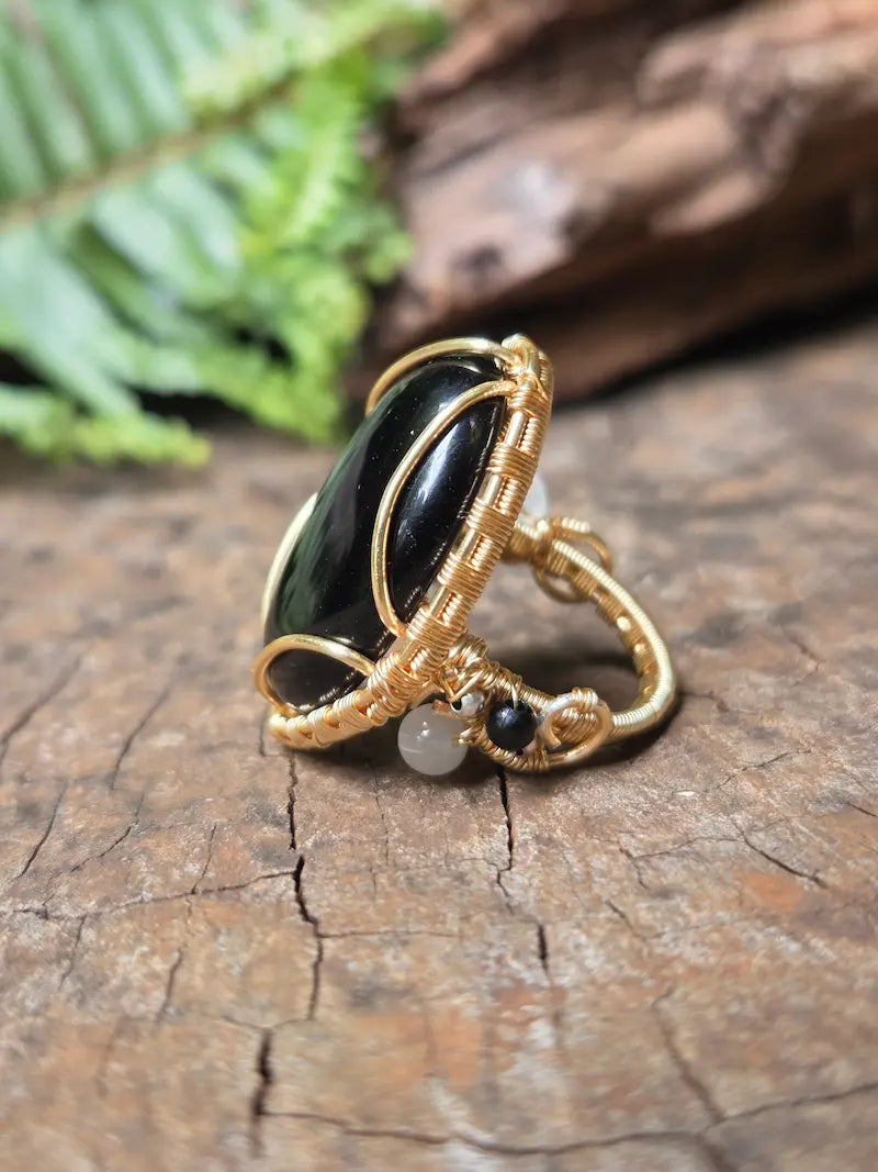 A copper wire-wrapped ring featuring a large black Obsidian cabochon and a small white bead, resting on a textured wooden surface with a blurred fern in the background.