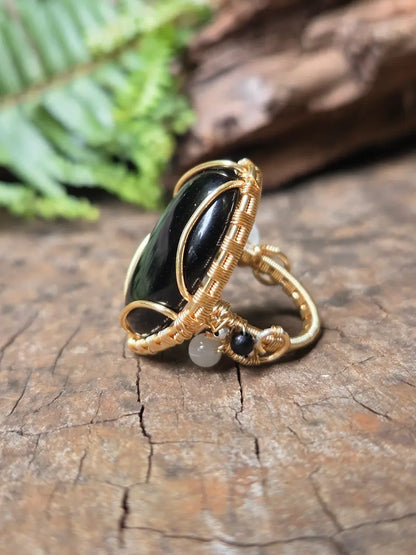 A copper wire-wrapped ring featuring a large black Obsidian cabochon and a small white bead, resting on a textured wooden surface with a blurred fern in the background.