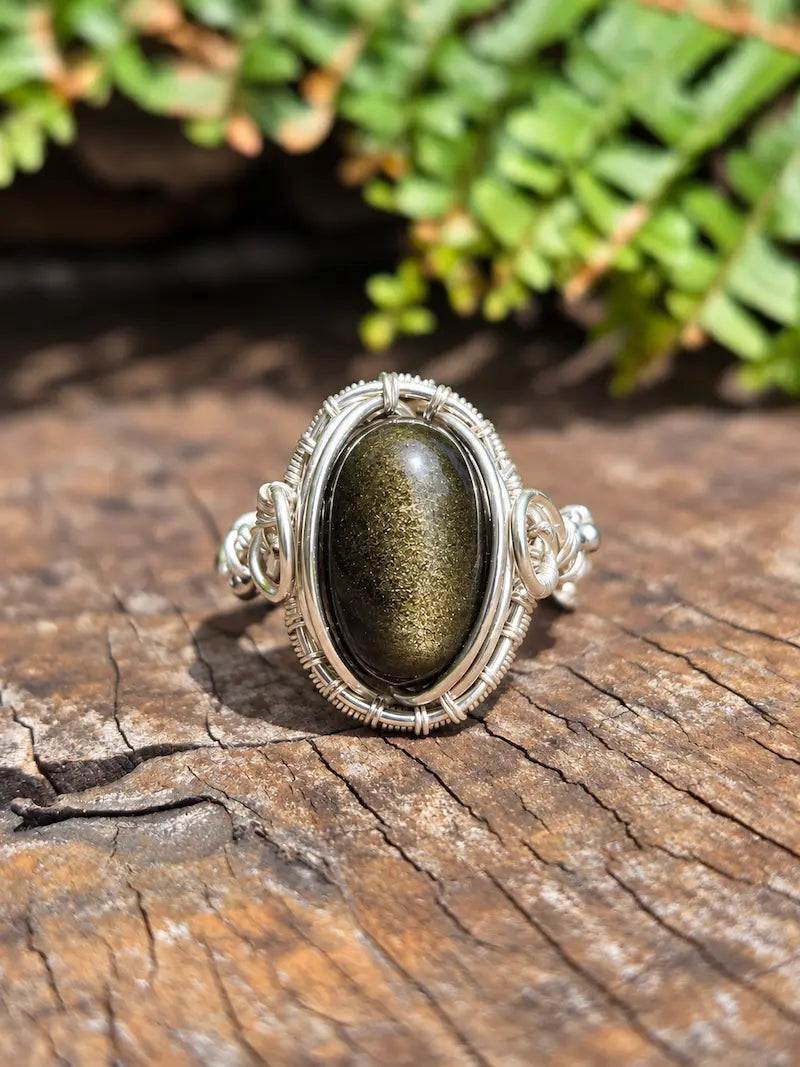 A handmade silver copper ring featuring an oval Black Obsidian stone with a gold sheen, set in a detailed silver bezel and wrapped with silver wire on a rustic wooden surface with green foliage in the background.
