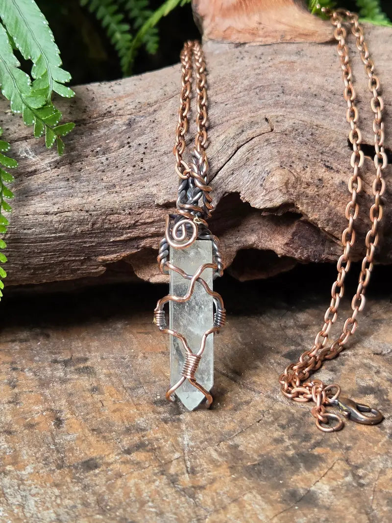 A handmade wire-wrapped crystal pendant necklace with a copper chain, displayed on a rustic piece of wood with green fern leaves in the background.