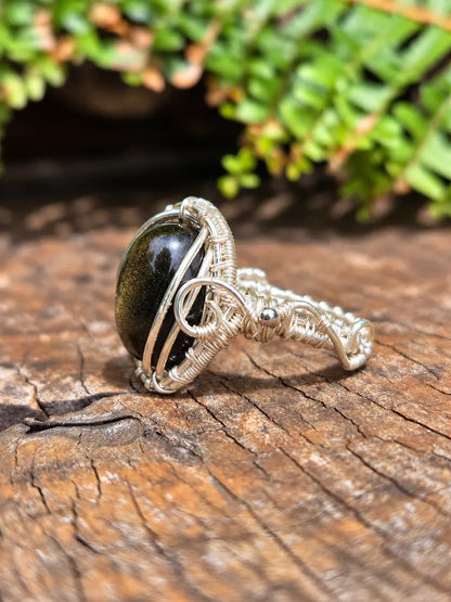 A handmade copper wire-wrapped ring featuring a large, polished black stone with subtle gold flecks, resting on a textured wooden surface with green foliage in the background.