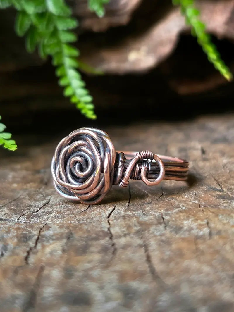 A handmade copper wire ring shaped like a spiral rose, resting on a textured wooden surface with green ferns in the background.