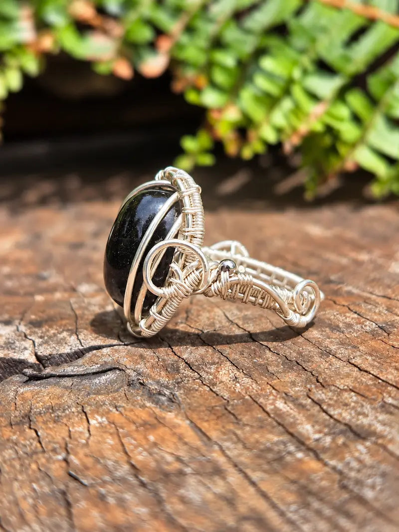 A handmade silver copper wire-wrapped ring featuring a smooth, oval black stone, resting on a textured wooden surface with a blurred green fern in the background.
