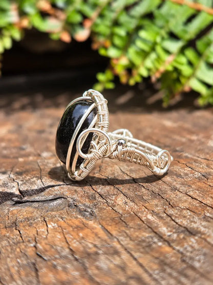 A handmade silver copper wire-wrapped ring featuring a smooth, oval black stone, resting on a textured wooden surface with a blurred green fern in the background.