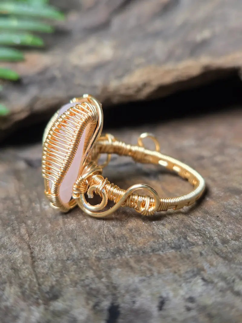 A handmade gold copper wire wrap ring featuring a pale pink stone, resting on a textured dark wood surface.