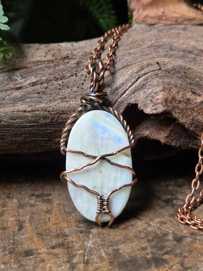 An oval pendant necklace featuring a white iridescent stone wrapped in copper wire, hanging from a matching copper chain on a rustic wood background.