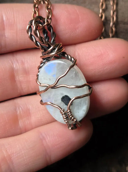 A close-up of a handmade pendant necklace featuring a raw, polished oval moonstone with a black inclusions and subtle blue flash, wrapped in copper wire, held in a person's hand.