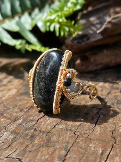 A handmade copper ring featuring a large, polished black stone wrapped in textured gold wire, resting on a rustic wooden surface with green ferns in the background.