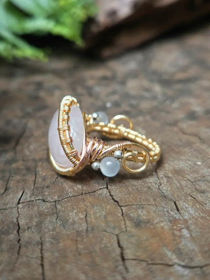 A handmade wire-wrapped ring featuring a large oval pale pink stone, a small round white bead, and tiny silver beads, set against a rustic wood background.