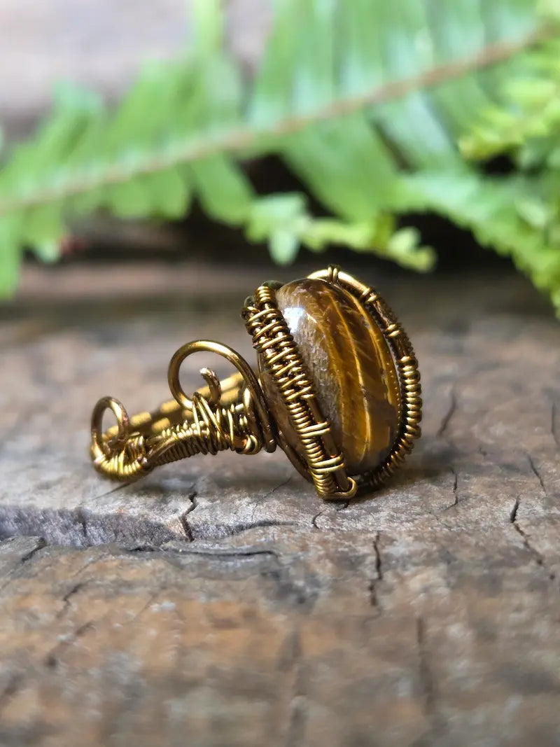 A handmade copper wire-wrapped ring featuring a polished tiger's eye stone, resting on a textured wooden surface with a blurred green fern in the background.