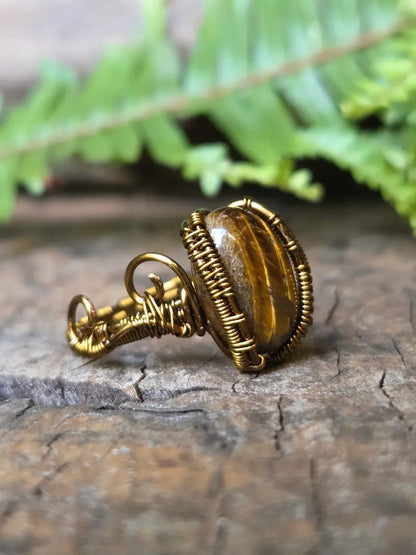 A handmade copper wire-wrapped ring featuring a polished tiger's eye stone, resting on a textured wooden surface with a blurred green fern in the background.