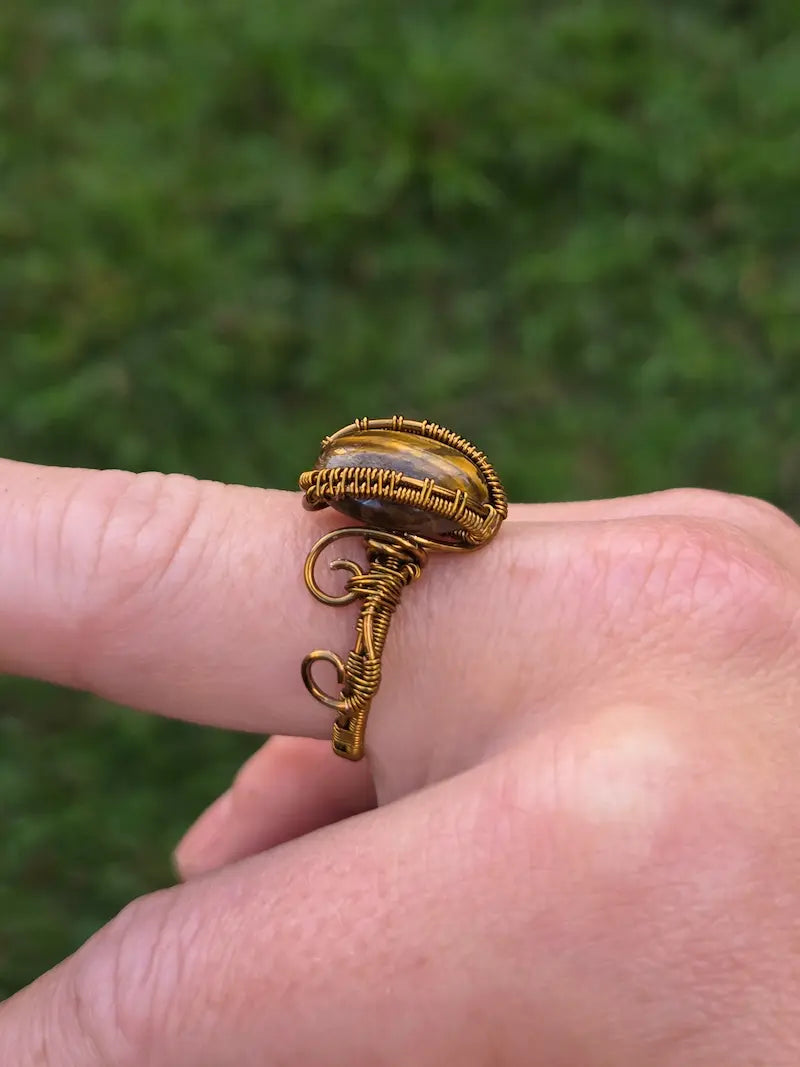 A close-up of a hand wearing a handmade copper wire wrap ring with a small oval stone, set against a blurred green outdoor background.