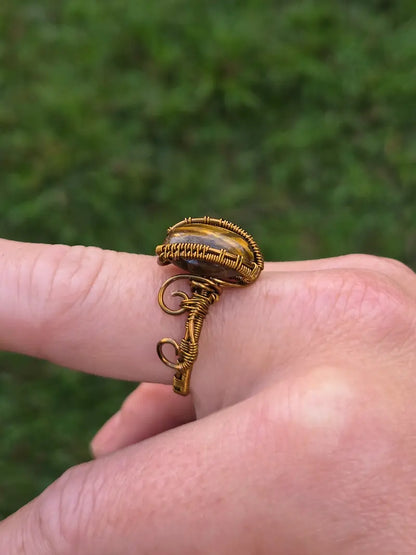 A close-up of a hand wearing a handmade copper wire wrap ring with a small oval stone, set against a blurred green outdoor background.
