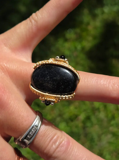 A close-up of a hand wearing a large ring with a black oval stone set in gold-toned copper metal, against a blurred green outdoor background.