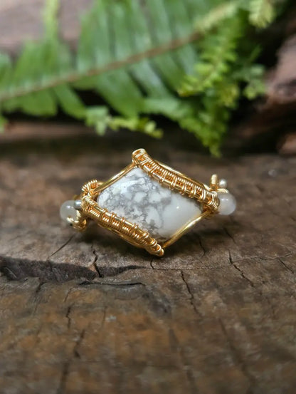 A handmade copper wire-wrapped gemstone pendant featuring a white howlite stone, resting on a textured wooden surface with a blurred green fern in the background.