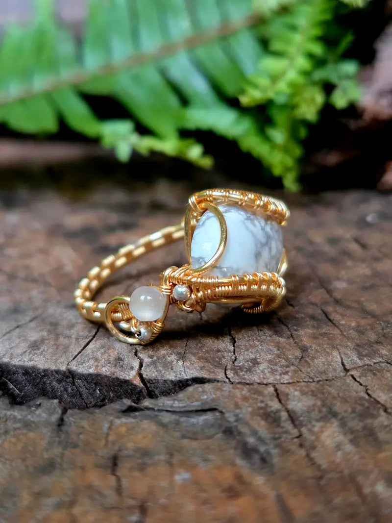 A handmade copper wire-wrapped gemstone pendant featuring a white howlite stone, resting on a textured wooden surface with a blurred green fern in the background.