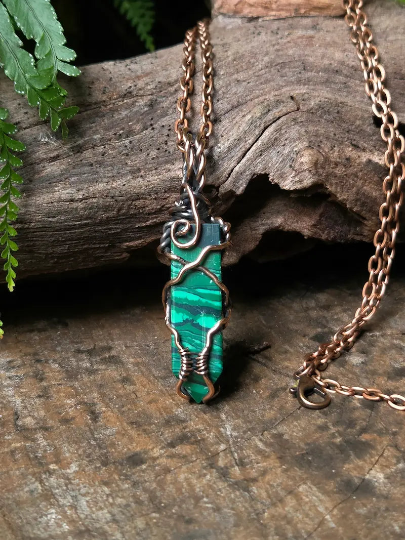 A handmade pendant necklace featuring a polished green malachite stone wrapped in copper wire, hanging from a copper chain on a rustic wood background.