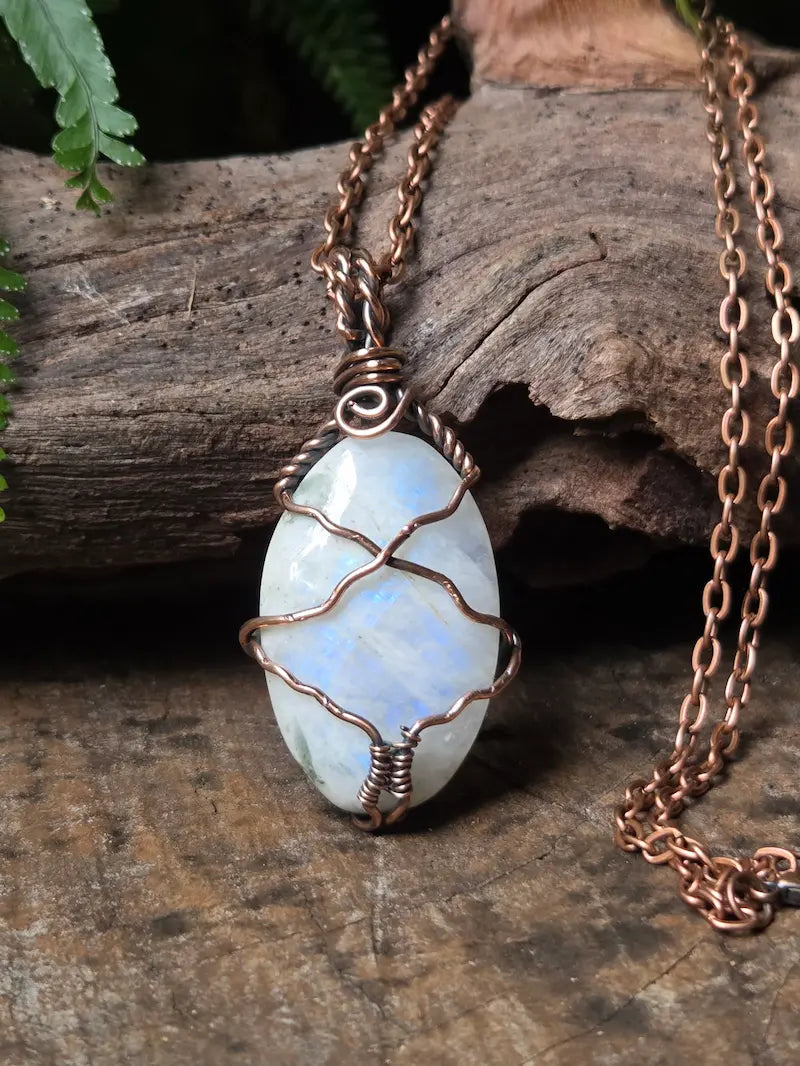 A copper wire-wrapped pendant necklace featuring an oval light blue and white moonstone, displayed on a rustic piece of wood with green leaves in the background.
