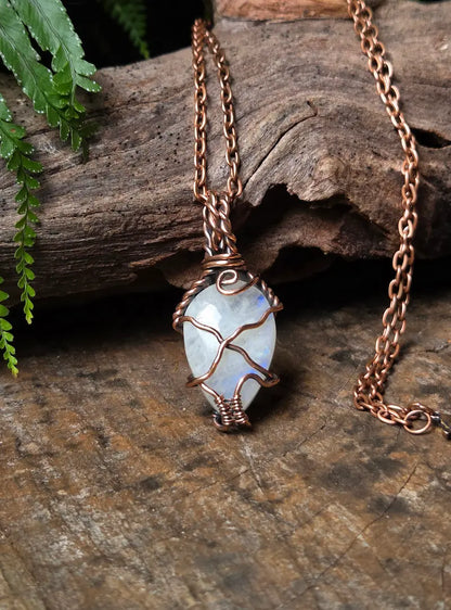 A copper wire-wrapped pendant necklace featuring a teardrop-shaped iridescent white gemstone, displayed on a rustic wooden surface with green leaves in the background.
