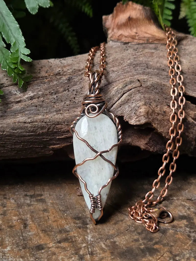 A handmade wire-wrapped pendant necklace featuring a white moonstone, displayed on a rustic piece of wood with green foliage in the background.