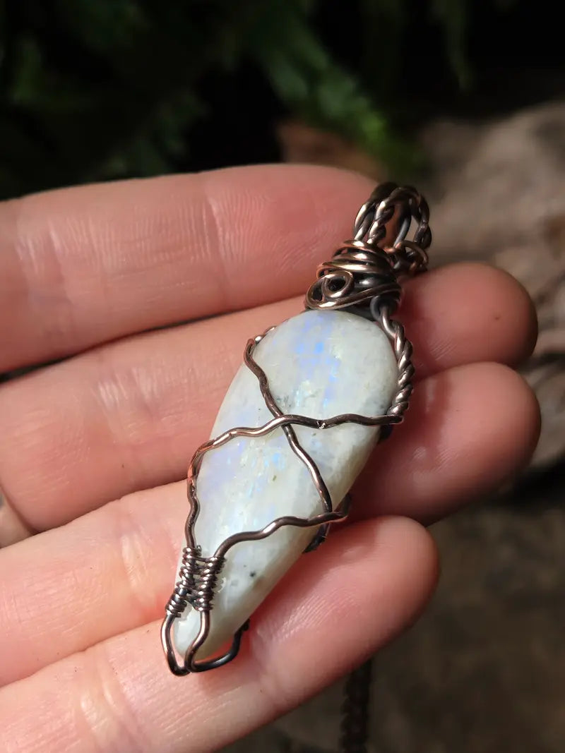 A hand holding a teardrop-shaped white moonstone pendant wrapped in textured copper wire, with a dark metal bail at the top.