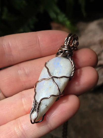 A hand holding a teardrop-shaped white moonstone pendant wrapped in textured copper wire, with a dark metal bail at the top.