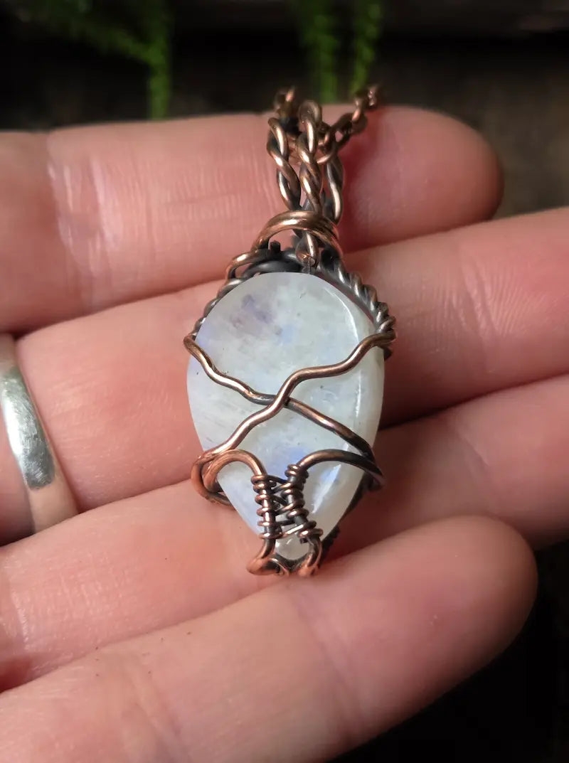 A handmade pendant featuring a teardrop-shaped white moonstone wrapped in copper wire, held in a person's hand.