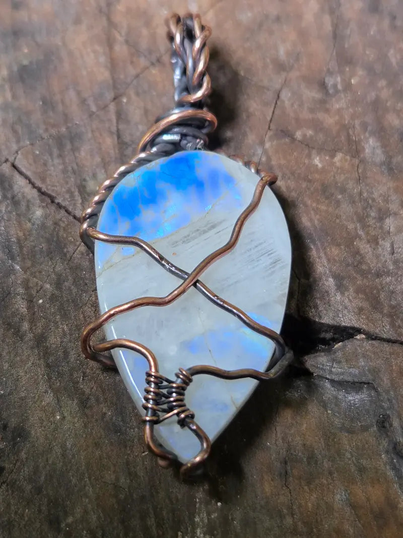 A teardrop-shaped moonstone pendant with a blue and gray play-of-color stone, wrapped in copper wire, resting on a textured dark wood surface.