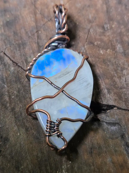 A teardrop-shaped moonstone pendant with a blue and gray play-of-color stone, wrapped in copper wire, resting on a textured dark wood surface.