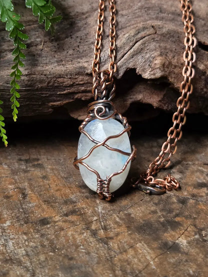 A handmade wire-wrapped necklace featuring an oval light blue-gray moonstone, displayed on a rustic wood background with a fern leaf in the corner.