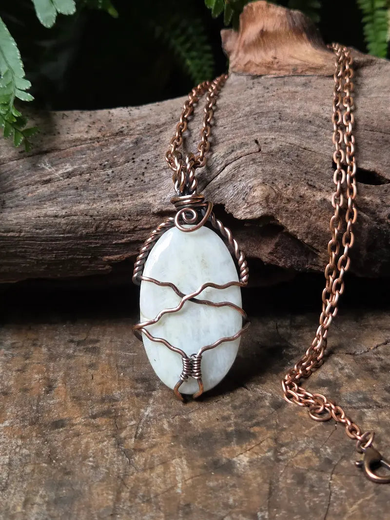 A handmade oval pendant necklace featuring a white stone wrapped in copper wire, resting on a piece of weathered wood with green leaves in the background.