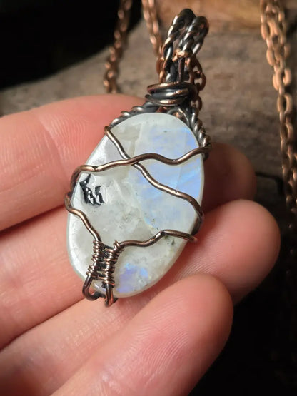 A close-up of a hand holding a moonstone pendant wrapped in copper wire, with a copper-toned chain visible in the background.