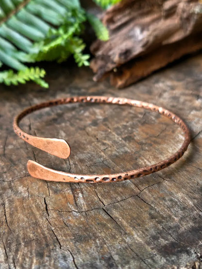 A hammered copper open cuff bracelet resting on a textured wooden surface with green ferns and driftwood in the background.