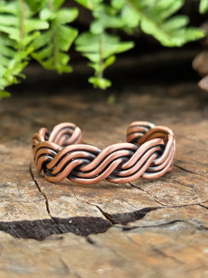 A habdmade copper twisted ring with darkened recesses, resting on a textured wooden surface with green fern leaves in the background.