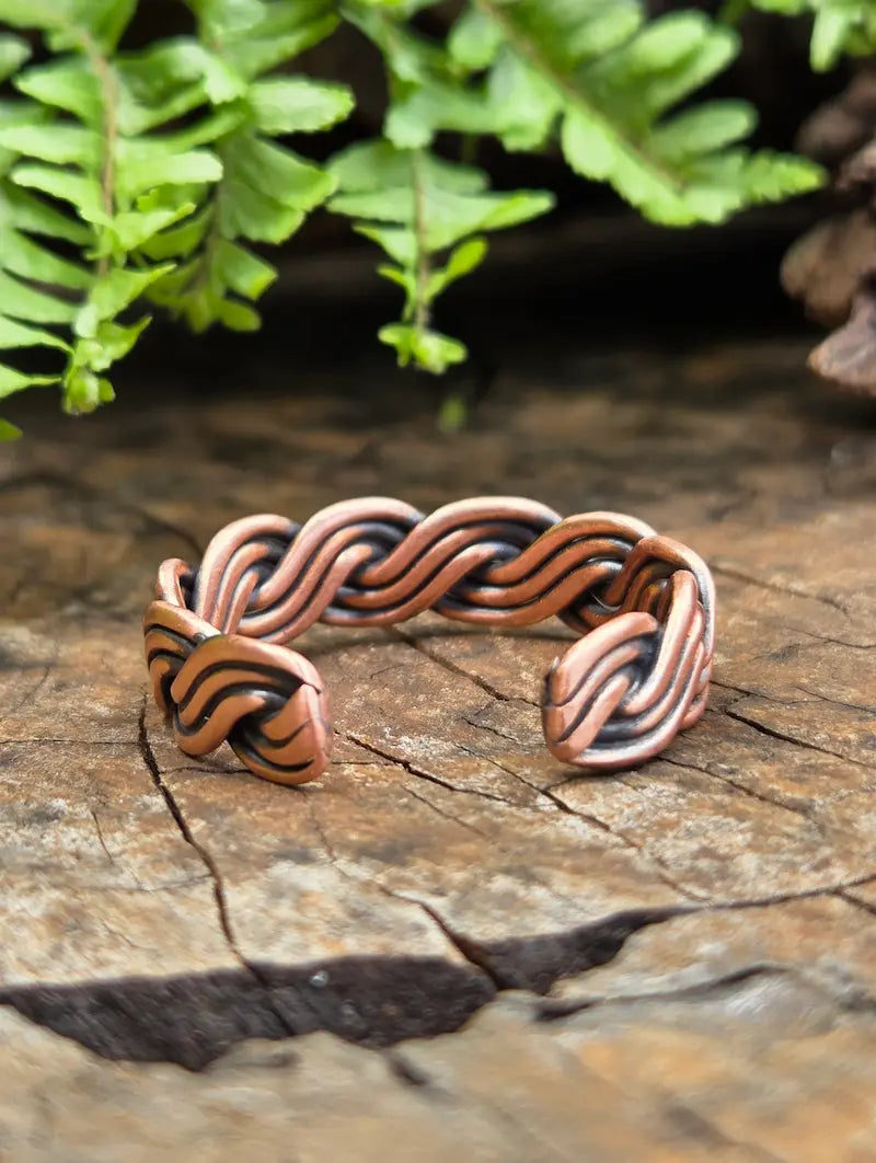 A handmade copper ring with a braided knot design, resting on a rustic wooden surface with green ferns in the background.