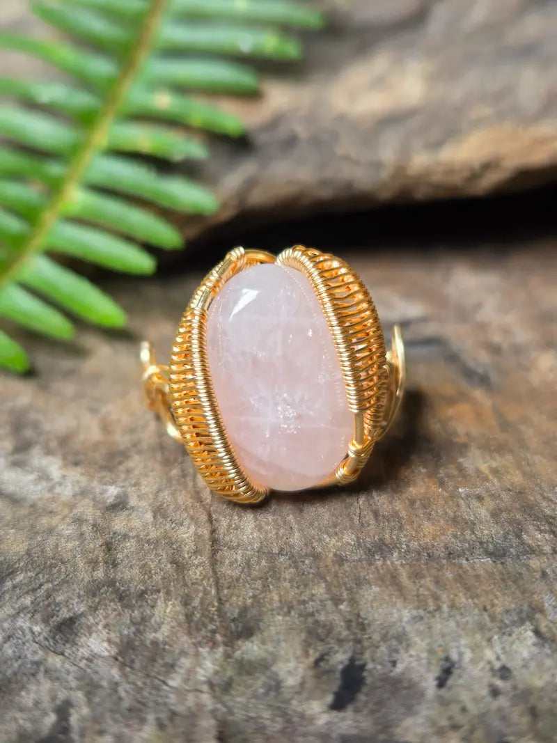 A gold copper ring with a large oval pale pink stone, photographed on a textured brown wood surface with a green fern leaf in the background.