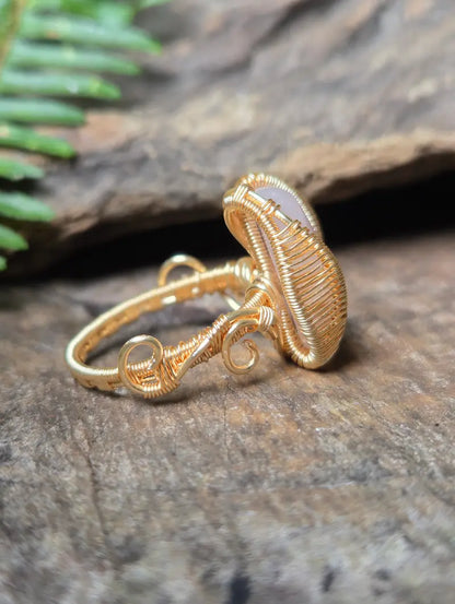 A copper wire-wrapped ring with a small oval pale pink rose quartz gemstone, resting on a textured brown wood surface with green leaves in the background.