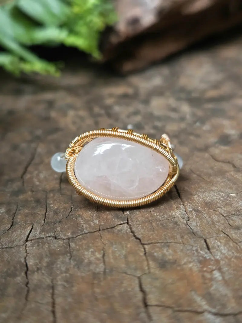 A handmade gold copper wire-wrapped ring featuring a rose quartz pink crystal and a small round white accent bead, resting on a natural wooden surface.