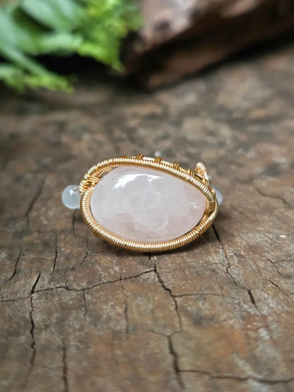A handmade gold copper wire-wrapped ring featuring a rose quartz pink crystal and a small round white accent bead, resting on a natural wooden surface.