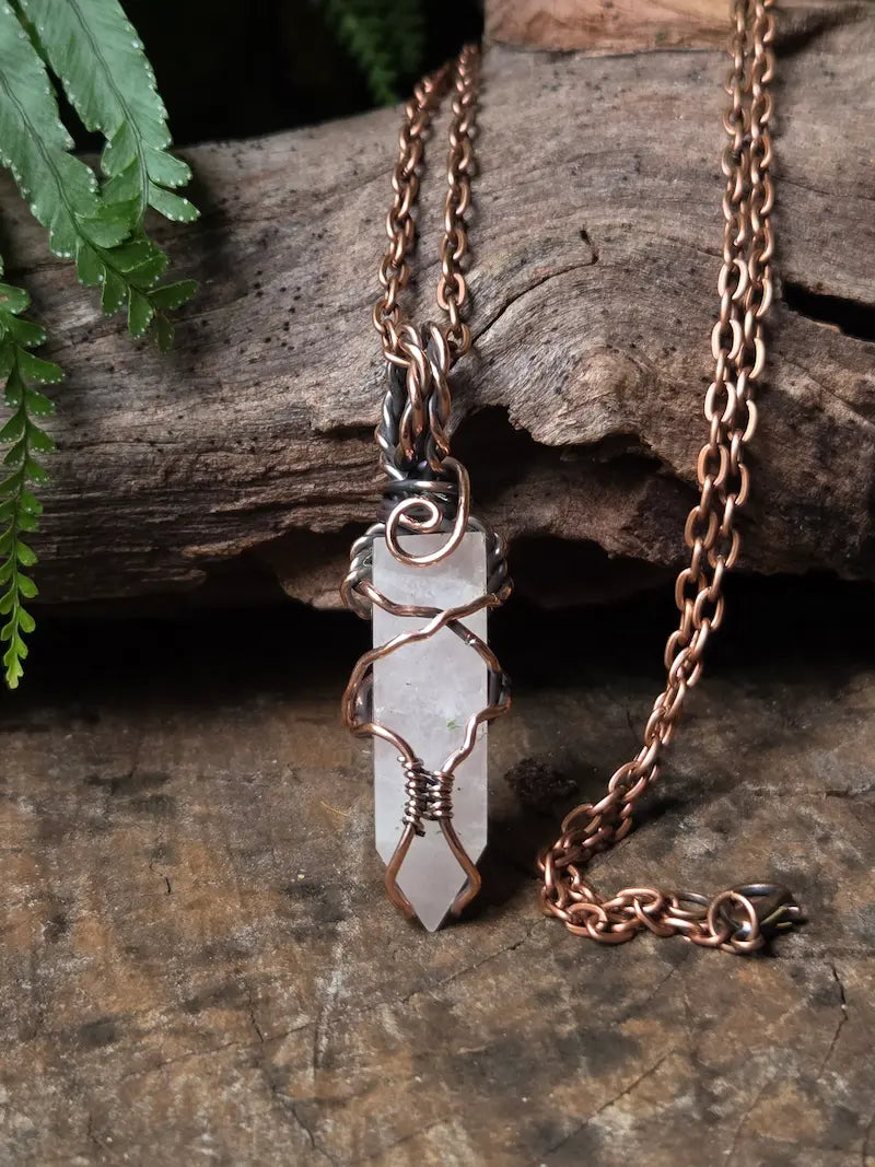 A handmade pendant necklace featuring a rose quartz point wrapped in copper wire, hanging from a matching copper chain on a rustic wood background.