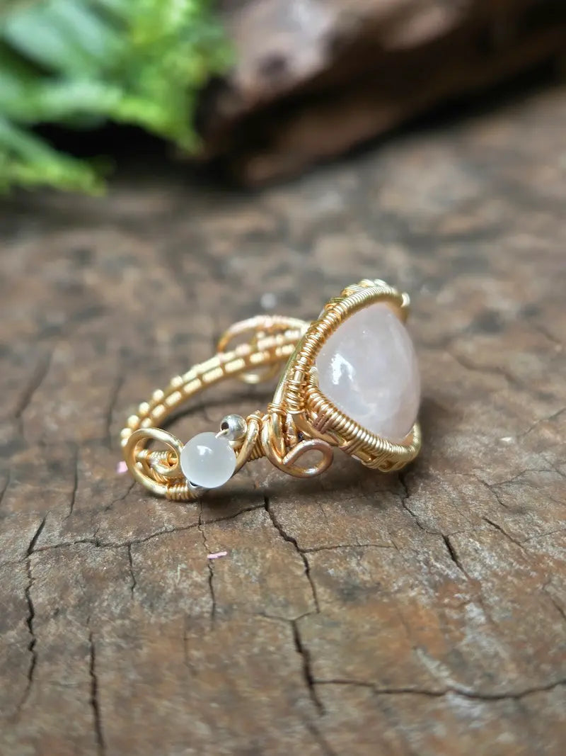 A handmade gold copper wire-wrapped ring featuring a rose quartz pink crystal and a small round white accent bead, resting on a natural wooden surface.