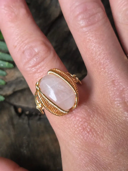 A gold copper wire-wrapped ring featuring a rose quartz pink stone, shown worn on a person's finger against a dark, rustic background.